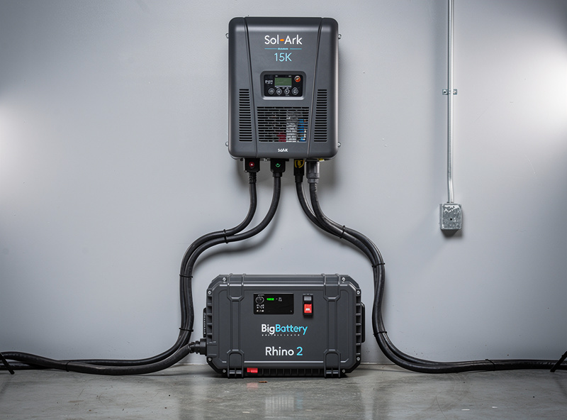 Sol-Ark 15K hybrid inverter mounted on a wall connected via cables to a BigBattery Rhino 2 freestanding battery unit on the floor below in a workshop