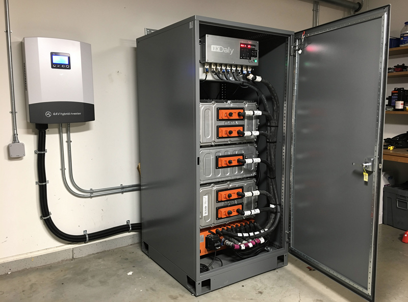 Used Tesla Model S battery modules installed in a fire rated enclosure with a Daly BMS and 24V hybrid inverter visible alongside in a workshop