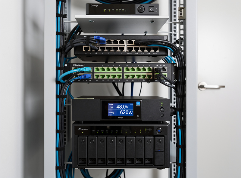 Wall-mounted homelab rack with router switch NAS server and battery management system display showing 48V state of charge and load wattage